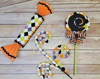 May include: Halloween themed decorations including a candy cane shaped lollipop with a black and white swirl design, a candy-shaped pillow with a black, white, orange, and yellow diamond pattern, and a black, white, orange, and yellow fabric swirl.
