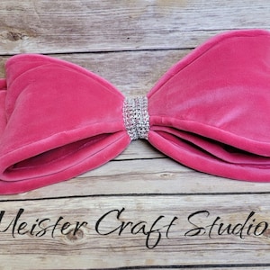 May include: A large pink velvet bow with a silver rhinestone center. The bow is lying on a wooden surface.