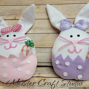 May include: Two plush Easter bunny decorations, one with a pink egg and the other with a purple egg. Both bunnies have white fur, pink noses, and black eyes. They are holding carrots in their paws.