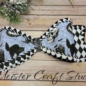 May include: A black and white bow with a Halloween theme. The bow is made of fabric with a black and white checkered pattern on the outside and a blue background with a black cat, skull, spider web, and other Halloween imagery on the inside. The bow is tied with a rhinestone embellishment.