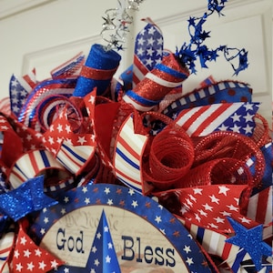 Op de afbeelding: Een rood-wit-blauwe patriottische krans met een stervormig middelpunt waarop "God Bless" staat. De krans is versierd met rood-wit-blauwe linten, gaas en sterren.