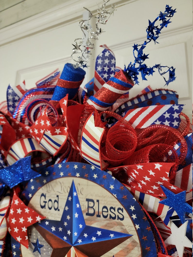 Op de afbeelding: Een rood-wit-blauwe patriottische krans met een stervormig middelpunt waarop "God Bless" staat. De krans is versierd met rood-wit-blauwe linten, gaas en sterren.