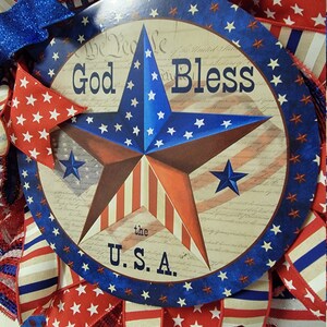 Op de afbeelding: Een rood-wit-blauwe patriottische krans met een rond bord waarop "God Bless the U.S.A." staat. Het bord toont een grote rood-wit-blauwe ster op een witte achtergrond en de tekst "We the People of the United States" op de achtergrond.