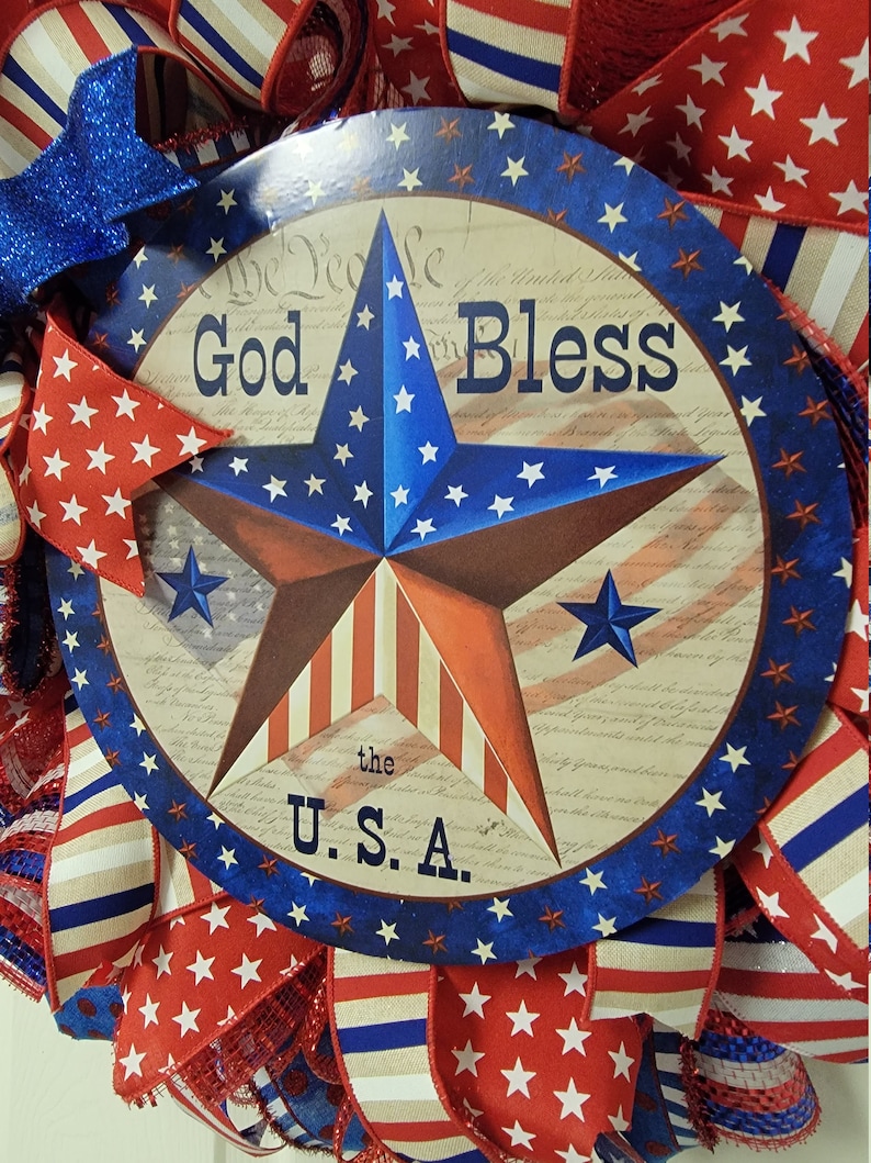 Op de afbeelding: Een rood-wit-blauwe patriottische krans met een rond bord waarop "God Bless the U.S.A." staat. Het bord toont een grote rood-wit-blauwe ster op een witte achtergrond en de tekst "We the People of the United States" op de achtergrond.