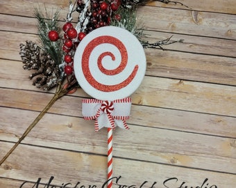 May include: A decorative Christmas lollipop with a red and white spiral design on a white background. The lollipop is adorned with a white ribbon and a red and white striped stick. The image also includes pine branches, red berries, and the text "Meister Craft Studio".