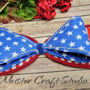 May include: A large, red, white, and blue bow with a star-spangled design. The bow is made of fabric and has a silver trim. The bow is on a wooden surface. Meister Craft Studio is written in black text at the bottom of the image.