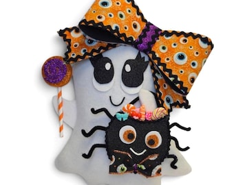 May include: A white felt ghost with a large orange bow adorned with eye patterns. The ghost holds a purple lollipop and a black spider with candy. The Halloween-themed decoration has black accents and is set against a white background.
