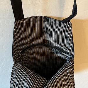 May include: A black crossbody bag with a black and white vertical striped fabric lining. The interior features a zippered pocket and a black nylon strap attached to the top. The bag is open, revealing the interior pattern and the zipper hardware.