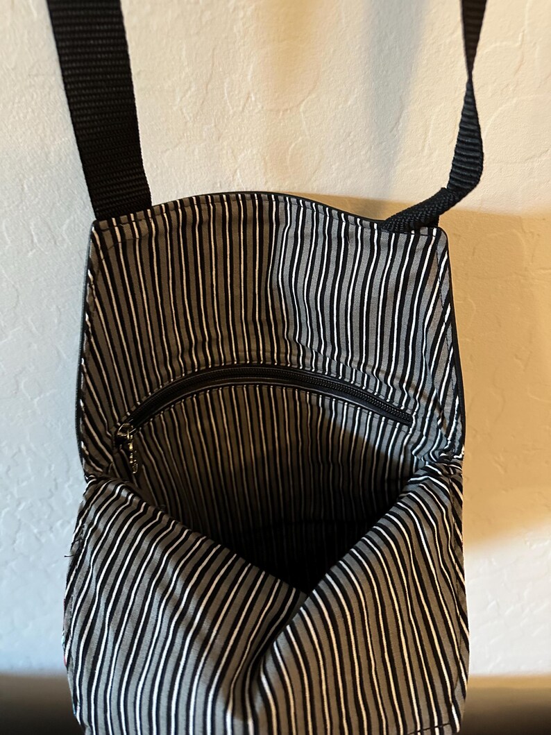 May include: A black crossbody bag with a black and white vertical striped fabric lining. The interior features a zippered pocket and a black nylon strap attached to the top. The bag is open, revealing the interior pattern and the zipper hardware.