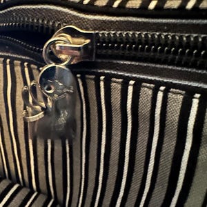 May include: A black and grey striped fabric bag featuring a silver-toned metal zipper closure. Attached to the zipper pull is a small, metallic charm shaped like a stylized character with a round head and thin limbs, finished in a polished silver tone.