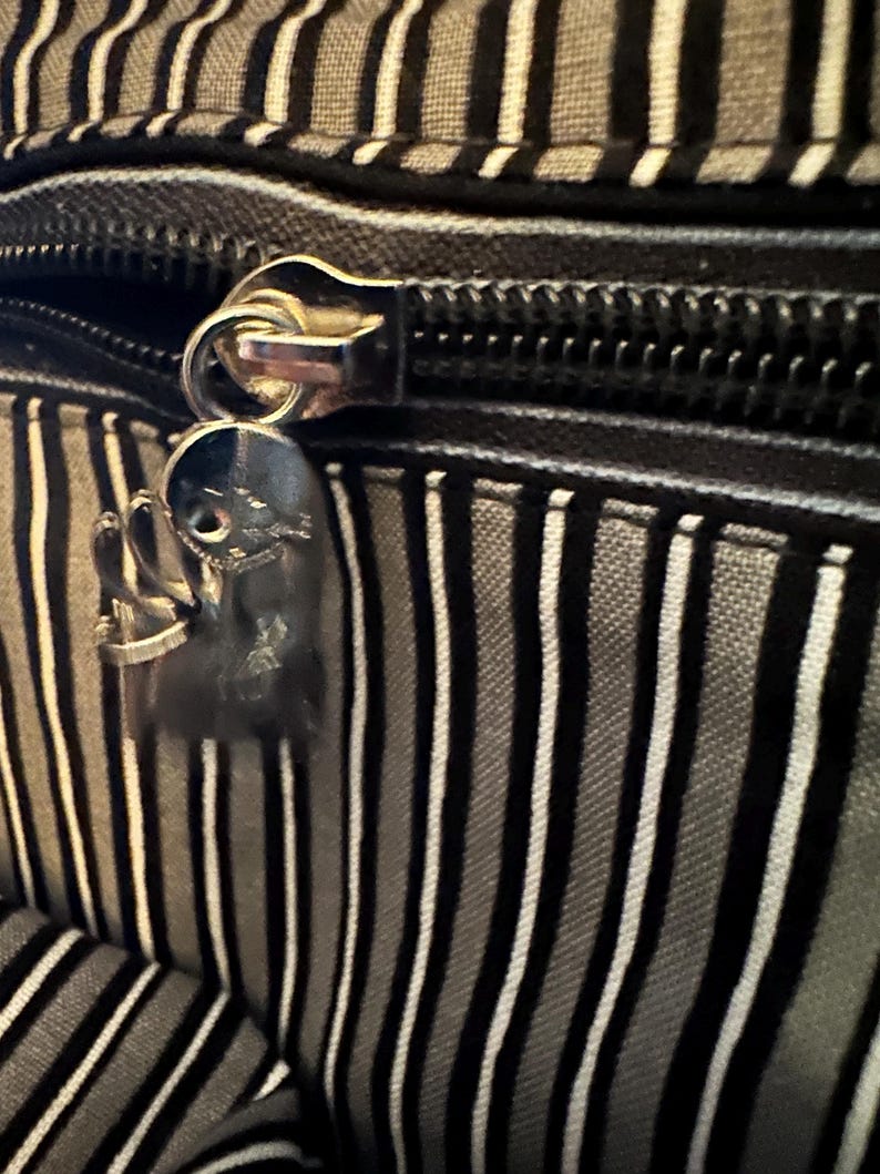 May include: A black and grey striped fabric bag featuring a silver-toned metal zipper closure. Attached to the zipper pull is a small, metallic charm shaped like a stylized character with a round head and thin limbs, finished in a polished silver tone.