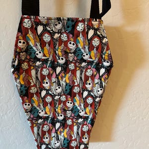May include: A coffin-shaped crossbody bag featuring a repeating pattern of Jack Skellington and Sally from The Nightmare Before Christmas. The bag has a black fabric exterior with a colorful character print and two black nylon shoulder straps.