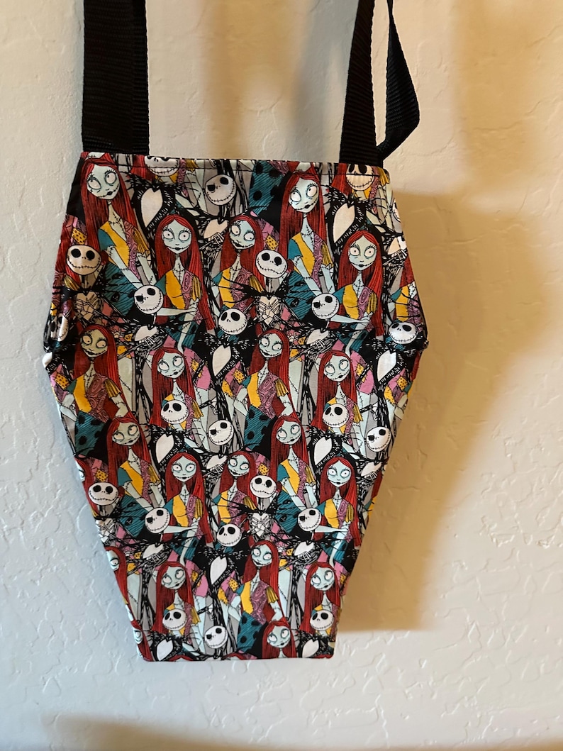 May include: A coffin-shaped crossbody bag featuring a repeating pattern of Jack Skellington and Sally from The Nightmare Before Christmas. The bag has a black fabric exterior with a colorful character print and two black nylon shoulder straps.