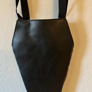 May include: A black coffin-shaped bag with a smooth, matte finish and a structured silhouette. The bag features a long, adjustable black nylon strap attached to the top, allowing it to be worn over the shoulder or across the body for convenience.