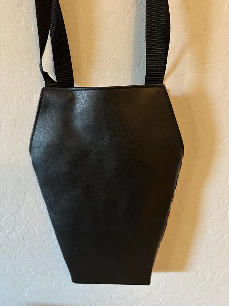 May include: A black coffin-shaped bag with a smooth, matte finish and a structured silhouette. The bag features a long, adjustable black nylon strap attached to the top, allowing it to be worn over the shoulder or across the body for convenience.