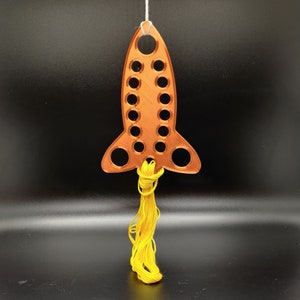 May include: A copper-colored rocket-shaped ornament with multiple circular cutouts. A yellow tassel is attached to the bottom, resembling flames. The ornament is suspended by a white string, suggesting it's designed for hanging. The background is a solid black.
