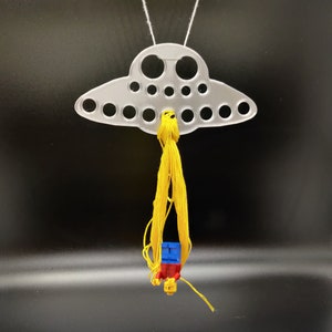 May include: A clear, UFO-shaped ornament with multiple circular cutouts, suspended by a white string. A yellow tassel hangs below, with a small Lego figure attached. The figure has a red torso, blue legs, and a yellow head.