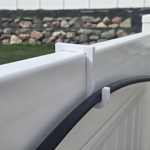 Può includere: Gancio bianco in plastica per recinzioni progettato per sostenere un tubo da giardino. Il gancio è attaccato a una recinzione in vinile bianco. Il tubo nero è parzialmente visibile, che si curva attraverso il gancio. Lo sfondo mostra un muro di pietra ed erba verde.