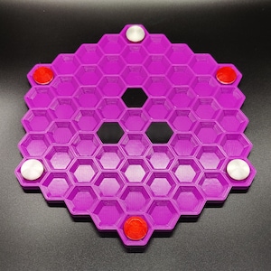 May include: A purple hexagonal board game with a honeycomb design. The board has several red and white circular game pieces. The game board is made of plastic and sits on a black surface.