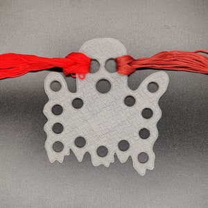 May include: A gray plastic ghost-shaped embroidery floss organizer with multiple holes. Red embroidery floss is threaded through the top holes. The organizer has a textured surface and a wavy bottom edge.