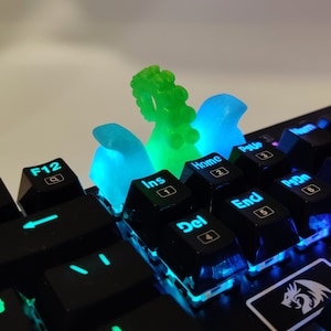 May include: A close-up of a black mechanical keyboard with custom keycaps. The keycaps are translucent and glow with blue and green light. The keycaps are shaped like tentacles.