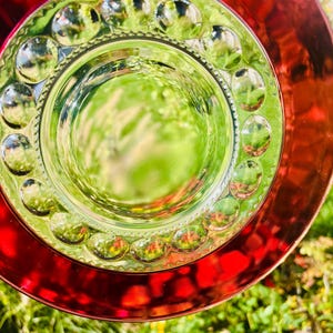 Puede incluir: Primer plano de un juego de platos decorativos de cristal. El plato presenta un borde exterior rojo, un borde interior verde con detalles circulares en relieve y un centro transparente. El plato refleja el follaje verde circundante.