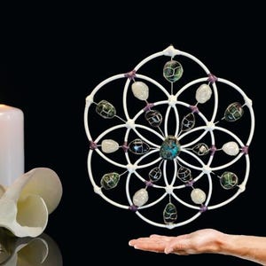 May include: A white, geometric wall hanging with a flower-like design. It features various gemstones in shades of green, white, and black, connected by a wire frame. The center stone is turquoise. The design is held by a hand.
