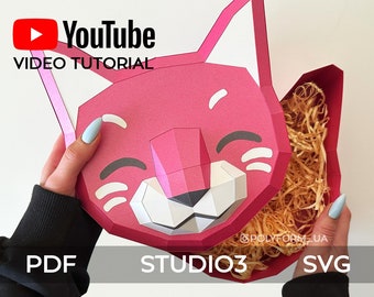 May include: A pink papercraft box shaped like a cartoon cat with a smiling face. The box is open and filled with shredded paper. The image includes the text "YouTube VIDEO TUTORIAL" and "STUDIO3".