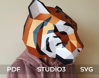 May include: A low-poly paper tiger mask featuring geometric facets in orange, white, and black. The mask displays the text PDF, STUDIO3, and SVG across the center. The mask is worn over a mauve hooded sweatshirt with visible drawstrings.
