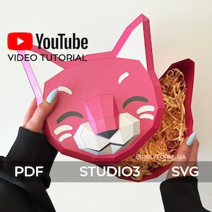 Puede incluir: Una caja de papel rosa con forma de gato de dibujos animados con una cara sonriente. La caja está abierta y llena de papel triturado. La imagen incluye el texto "YouTube VIDEO TUTORIAL" y "STUDIO3".