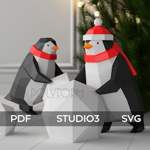 May include: Two origami penguins made of paper, one wearing a red Santa hat and scarf, are rolling a large white snowball. The penguins are standing on a white wooden surface. The text "PDF STUDIO3 SVG" is visible below the penguins.