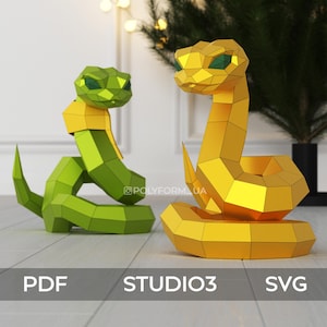 Puede incluir: Dos serpientes de papel, una verde y otra dorada, están colocadas en posición enroscada. Las serpientes tienen ojos verdes y están hechas de papel doblado. Las serpientes están sentadas sobre una superficie de madera.