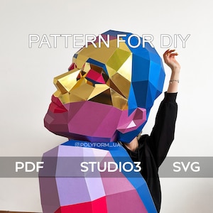 Puede incluir: Un modelo de papelcraft de cabeza humana colorido, con un esquema de color azul, dorado, rosa y morado. El modelo lo sostiene una persona que lleva una camisa negra y pantalones vaqueros. El texto "PATTERN FOR DIY" está en la parte superior de la imagen, y el texto "PDF STUDIO3 SVG" está en la parte inferior de la imagen.