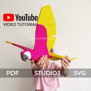 May include: A pink and yellow papercraft parrot with a YouTube logo and the text "VIDEO TUTORIAL" and "@POLYFORM_UA". The parrot is being held by a person. The text "PDF", "STUDIO3", and "SVG" is also visible.