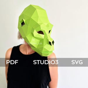 May include: A green papercraft gorilla mask. The mask is made of folded paper and has a realistic design. The mask is being worn by a person with black clothing. The text "STUDIO3" is visible in the image.