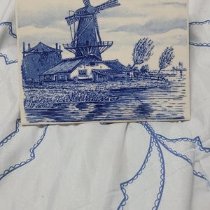 以下が含まれることがあります： 青と白のセラミックタイルに、手描きのオランダの風車と家の絵が描かれています。風車は白い本体と青い羽根を持っています。家は白で青い縁取りがされています。タイルは青い縁取りがされています。