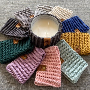K&ouml;nnte beinhalten: Eine Sammlung geh&auml;kelter Kerzenw&auml;rmer in verschiedenen Farben, darunter Gr&uuml;n, Blau, Rosa, Lila und Gelb. Die W&auml;rmer sind um eine wei&szlig;e Kerze in einem Glasgef&auml;&szlig; angeordnet.