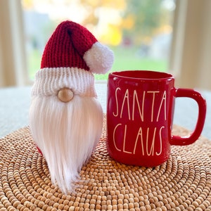 Könnte beinhalten: Ein rot-weißer Häkel-Zwerg mit einem Weihnachtsmann-Hut sitzt neben einer roten Tasse mit der Aufschrift "Santa Claus". Die Tasse und der Zwerg stehen auf einem geflochtenen Platzdeckchen.