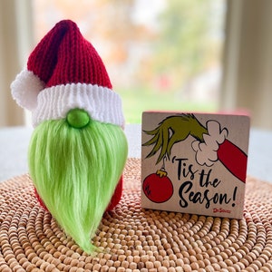 Könnte beinhalten: Ein Weihnachtszwerg mit roter Strickmütze und grünem Bart. Der Zwerg hat einen weißen Strickrand und eine grüne Nase. Neben dem Zwerg befindet sich ein Holzblock mit dem Text "'Tis the Season!" und einer Illustration von Dr. Seuss.