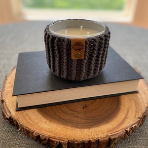 K&ouml;nnte beinhalten: Ein grauer, geh&auml;kelter Kerzenhalter mit einem braunen Leder-Etikett mit der Aufschrift "Made with Love" steht auf einem schwarzen Buch, das auf einer Holzscheibe liegt.