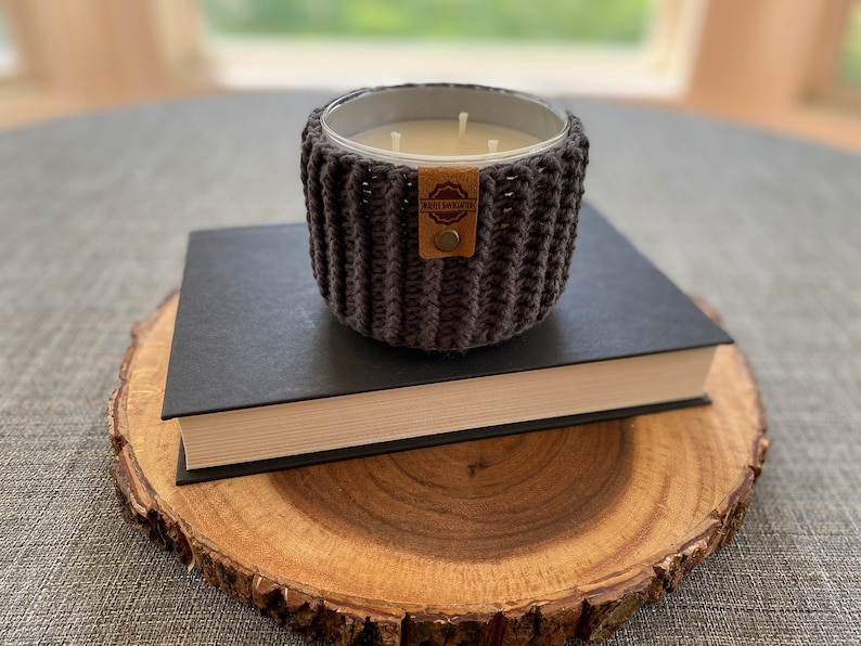 K&ouml;nnte beinhalten: Ein grauer, geh&auml;kelter Kerzenhalter mit einem braunen Leder-Etikett mit der Aufschrift "Made with Love" steht auf einem schwarzen Buch, das auf einer Holzscheibe liegt.