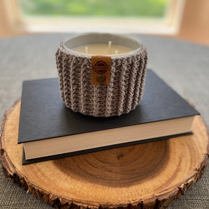 K&ouml;nnte beinhalten: Ein grauer geh&auml;kelter Kerzenhalter mit einem braunen Leder-Etikett, auf dem "Made with Love" steht. Der Kerzenhalter steht auf einem schwarzen Buch, das auf einer Holzscheibe liegt.