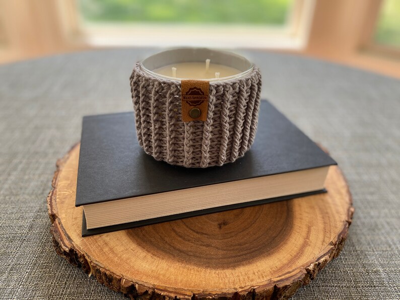 K&ouml;nnte beinhalten: Ein grauer geh&auml;kelter Kerzenhalter mit einem braunen Leder-Etikett, auf dem "Made with Love" steht. Der Kerzenhalter steht auf einem schwarzen Buch, das auf einer Holzscheibe liegt.
