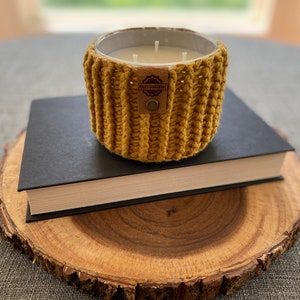 K&ouml;nnte beinhalten: Ein gelber geh&auml;kelter Kerzenhalter mit einem braunen Leder-Etikett mit der Aufschrift "Kati Hawthorn" steht auf einem schwarzen Buch, das auf einer Holzscheibe liegt.