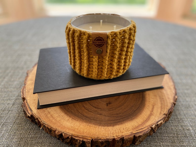K&ouml;nnte beinhalten: Ein gelber geh&auml;kelter Kerzenhalter mit einem braunen Leder-Etikett mit der Aufschrift "Kati Hawthorn" steht auf einem schwarzen Buch, das auf einer Holzscheibe liegt.