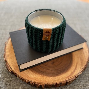 K&ouml;nnte beinhalten: Ein gr&uuml;nes, geh&auml;keltes Kerzengef&auml;&szlig; mit einer wei&szlig;en Kerze und einem braunen Etikett mit der Aufschrift "HAND CRAFTED". Das Kerzengef&auml;&szlig; steht auf einem schwarzen Buch auf einer Holzscheibe.