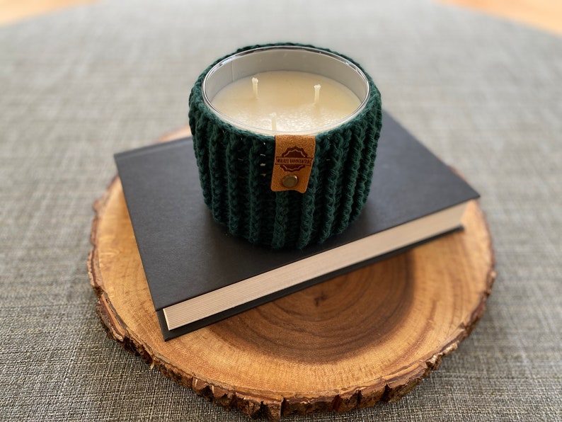 K&ouml;nnte beinhalten: Ein gr&uuml;nes, geh&auml;keltes Kerzengef&auml;&szlig; mit einer wei&szlig;en Kerze und einem braunen Etikett mit der Aufschrift "HAND CRAFTED". Das Kerzengef&auml;&szlig; steht auf einem schwarzen Buch auf einer Holzscheibe.