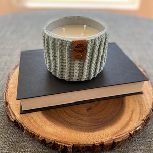 K&ouml;nnte beinhalten: Ein hellblaues, gestricktes Kerzengef&auml;&szlig; mit einem braunen Lederaufkleber mit der Aufschrift "Remarkable" auf einem schwarzen Buch und einer Holzscheibe.
