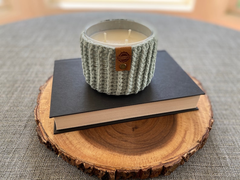K&ouml;nnte beinhalten: Ein hellblaues, gestricktes Kerzengef&auml;&szlig; mit einem braunen Lederaufkleber mit der Aufschrift "Remarkable" auf einem schwarzen Buch und einer Holzscheibe.