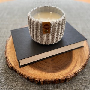 K&ouml;nnte beinhalten: Ein grauer gestrickter Kerzenhalter mit einem braunen Leder-Etikett mit der Aufschrift "Handmade with Love" steht auf einem schwarzen Buch, das auf einer Holzscheibe liegt.